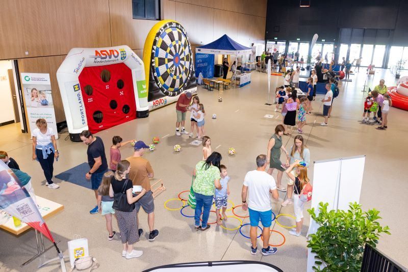 Familien und Kinder spielen mit Bällen und Darts bei einer Veranstaltung. Verschiedene aufblasbare Spiele sind im Raum aufgestellt.