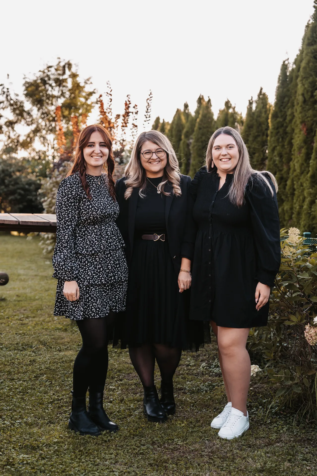 Drei Frauen stehen lächelnd und posieren für ein Foto in einem Garten. Sie sind von Pflanzen und Bäumen umgeben.
