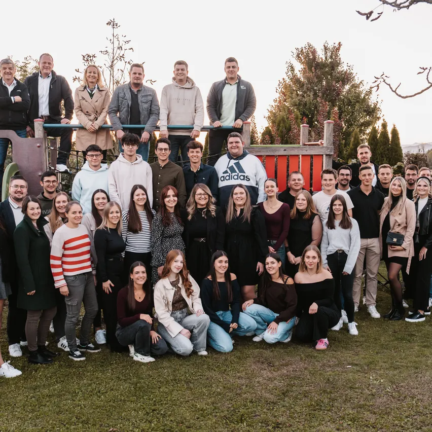 Eine Gruppe von Menschen posiert für ein Foto in einem Park, mit Bäumen und einer Spielstruktur im Hintergrund.