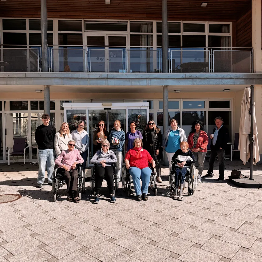Eine Gruppe von Menschen in Rollstühlen und im Stehen posiert für ein Foto vor einem Gebäude mit Balkon.