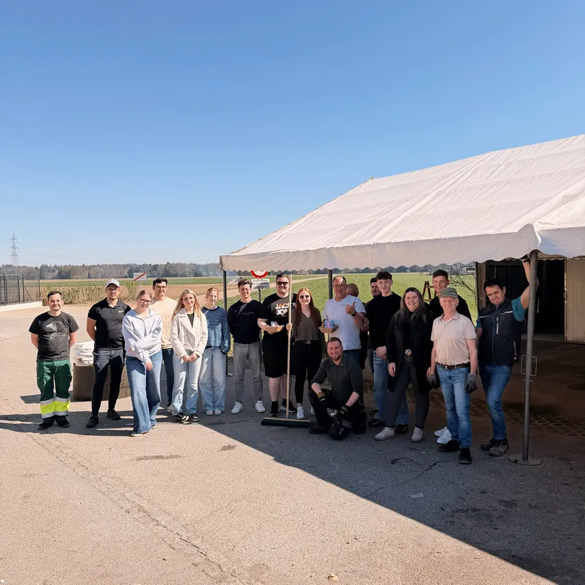 Eine Gruppe von Menschen posiert für ein Foto unter einem Zelt. Die Gruppe umfasst einen Mann, der fegt, und eine Frau, die einen Tablett hält. Sie sind in der Nähe einer Straße, mit einem Feld im Hintergrund.