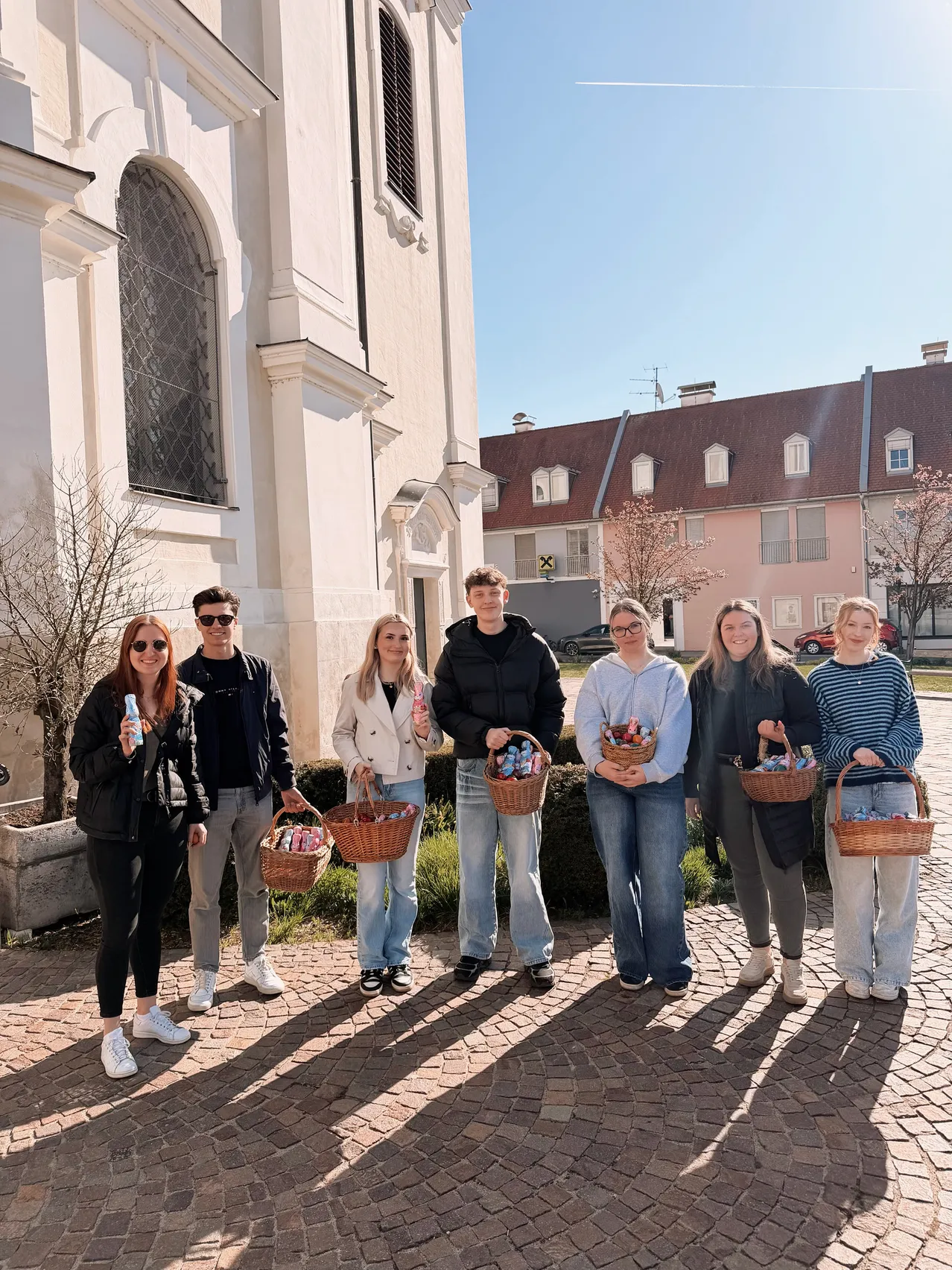 Sieben Personen stehen vor einer Kirche, jede hält Körbe mit Ostereiern. Sie sind in Winterkleidung gekleidet. Dahinter befindet sich ein Haus und ein Baum.