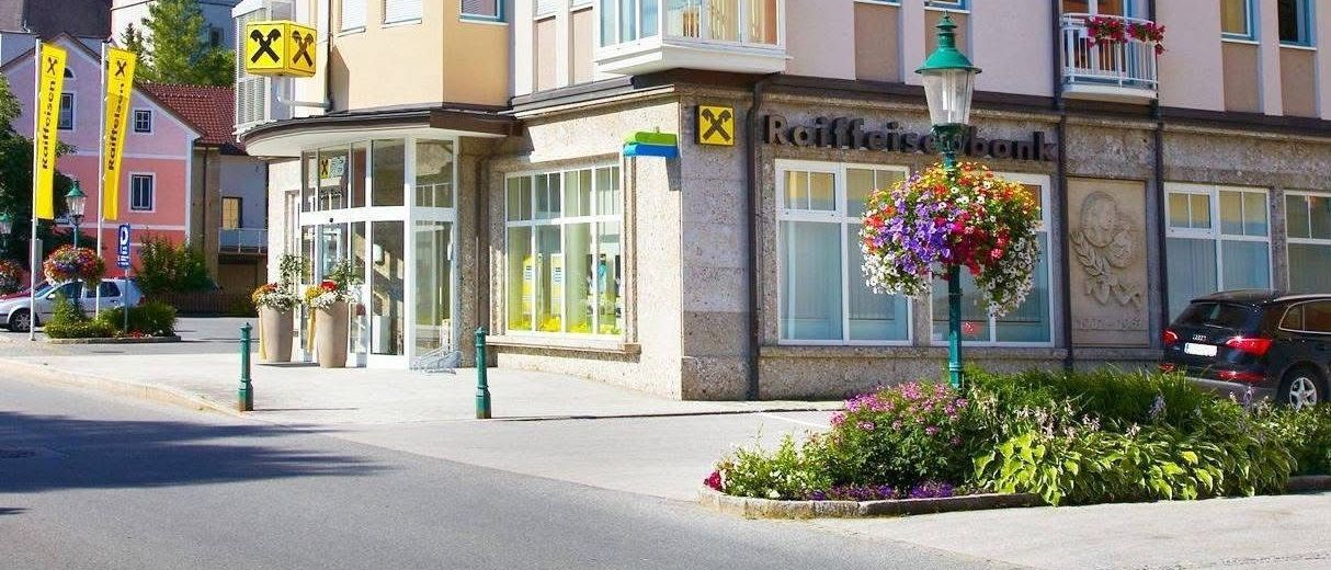 Eine Straßenszene mit einem Gebäude, das ein Raiffeisenbank-Schild trägt. Das Gebäude hat eine Mischung aus gelben und weißen Farben, mit blauen Fenstern und Balkonen. Im Hintergrund befindet sich ein Turm.
