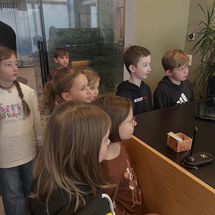 Mehrere Kinder stehen um einen Schreibtisch herum, schauen auf einen Computerbildschirm, mit einer Pflanze im Hintergrund.