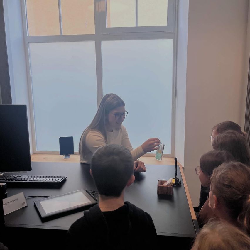 Eine Frau mit Brille zeigt einem Kindersitzkreis einen kleinen Behälter. Der Tisch vor ihnen hat einen Monitor, eine Tastatur und ein Tablet.