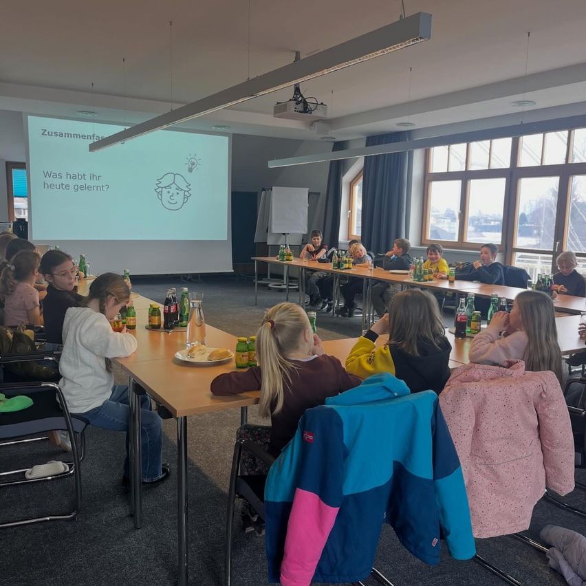 Eine Gruppe von Kindern sitzt in einem Klassenzimmer an Tischen, der Projektionsleinwand zugewandt. Einige von ihnen essen, während andere die Präsentation beobachten. Auf den Tischen liegen Flaschen, Teller und Jacken. Eine Lehrerin sitzt hinten.