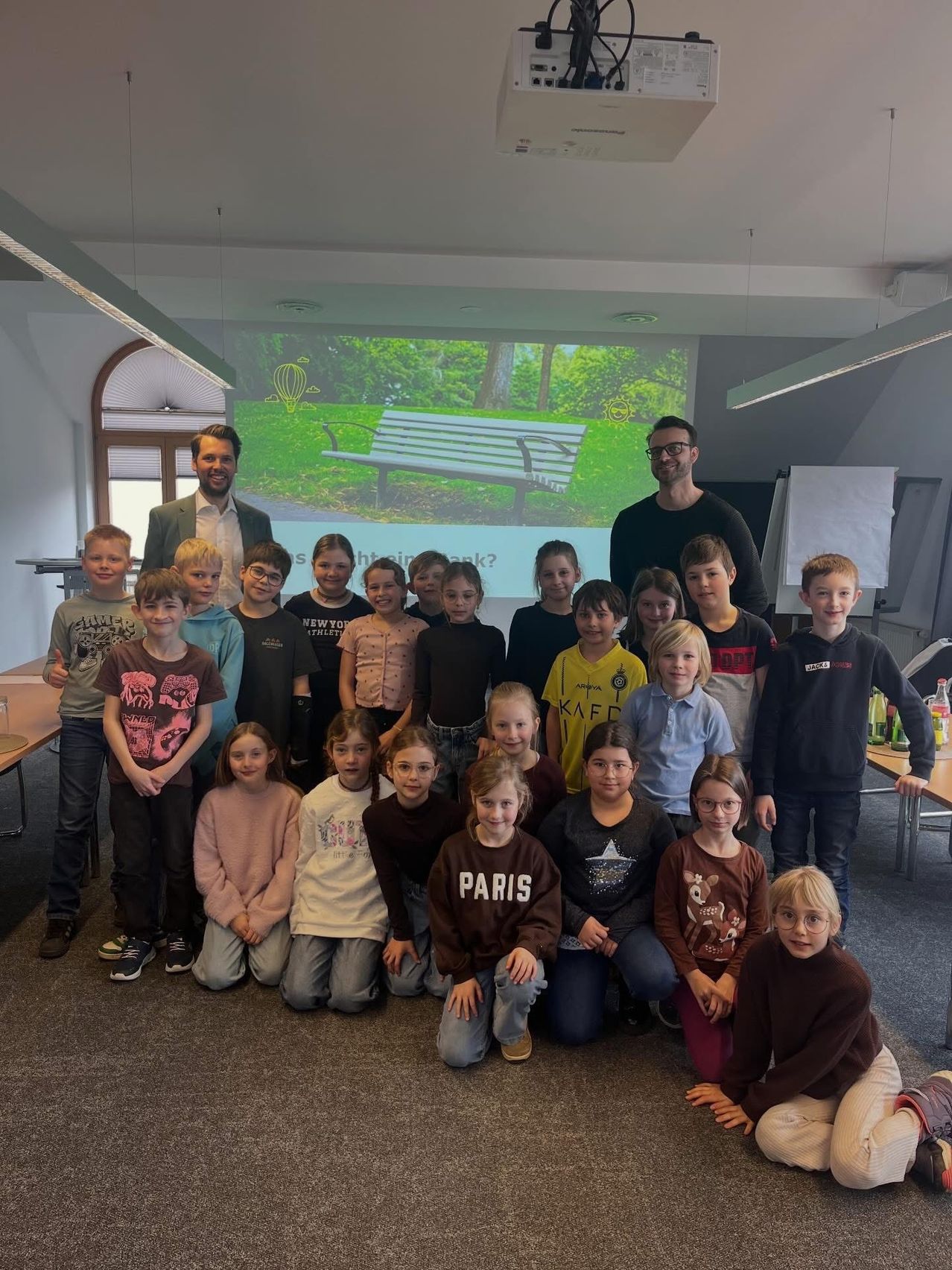 Eine Gruppe von Kindern und Erwachsenen steht vor einer Projektionsleinwand mit einer Parkbank. Die Kinder lächeln und posieren für ein Foto. Die Erwachsenen stehen hinter den Kindern.