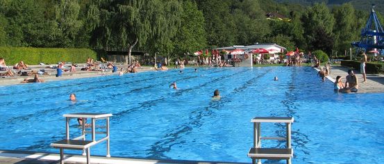 Ein großer Freiluft-Schwimmbad mit Schwimmern und anderen, die sich an den Seiten entspannen, umgeben von üppigem Grün und einem klaren blauen Himmel.