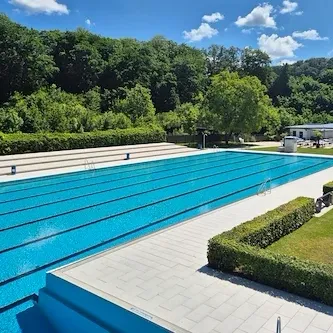 Ein Freibad mit klarem blauem Wasser, umgeben von üppigem Grün und einem klaren blauen Himmel mit einigen Wolken.