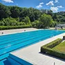 Ein Freibad mit klarem blauem Wasser, umgeben von üppigem Grün und einem klaren blauen Himmel mit einigen Wolken.