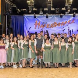 Eine Gruppe von Menschen in traditioneller bayerischer Kleidung, einschließlich grüner Kleider und Lederhosen, posiert für ein Foto auf einer Bühne mit Musikinstrumenten und einem Banner, das 'DIE HAFENDORFER' steht.
