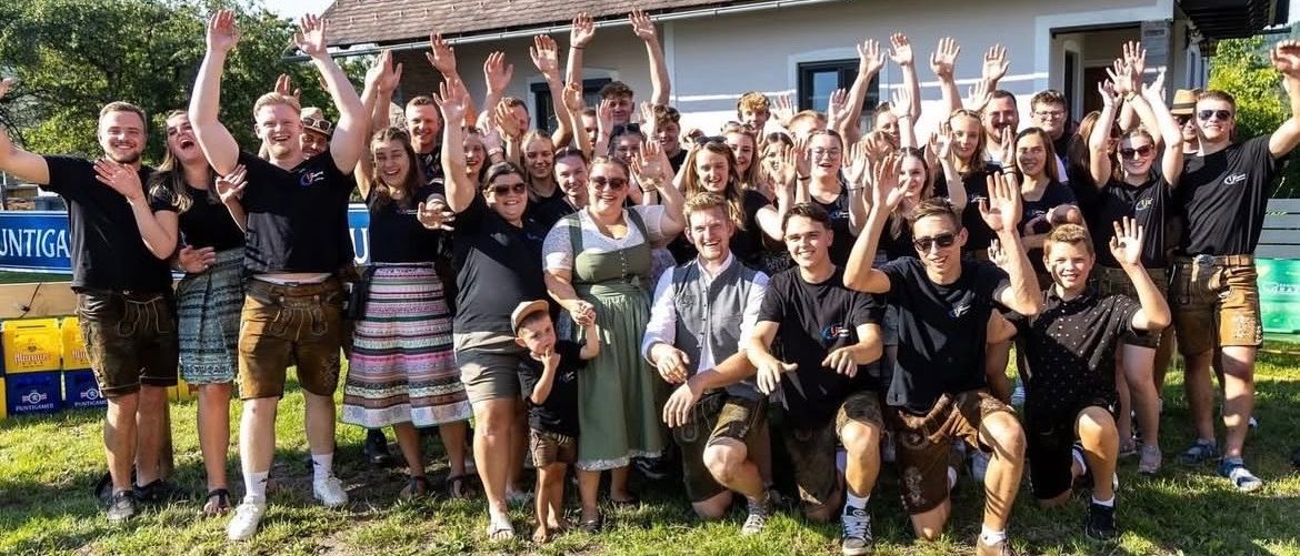 Eine Gruppe von Menschen steht vor einem Haus mit Solarpanelen, alle lächeln und heben ihre Hände. Einige tragen traditionelle Kleidung, andere Freizeitkleidung. Ein Kind wird von einem Erwachsenen gehalten.