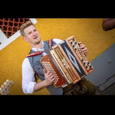 Ein Mann in einer Weste spielt Akkordeon. Er lächelt und hält das Instrument. Er steht vor einer gelben Wand.