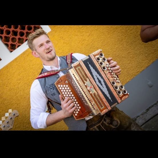 Ein Mann in einer Weste spielt Akkordeon. Er lächelt und hält das Instrument. Er steht vor einer gelben Wand.