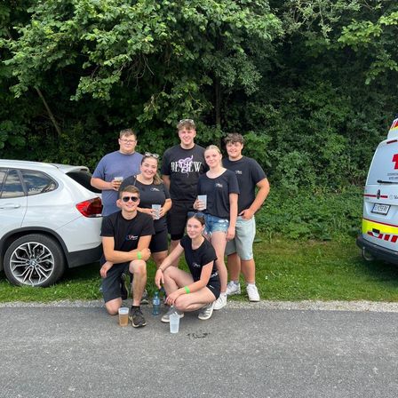 Eine Gruppe junger Menschen, die lächelnd für ein Foto posiert, steht am Straßenrand, mit einem Auto und einem Krankenwagen im Hintergrund.