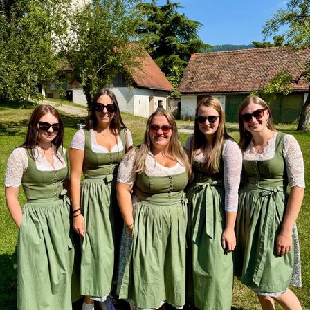 Fünf Frauen in grünen Kleidern stehen vor einem Haus und einem Baum an einem sonnigen Tag.