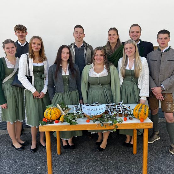 Eine Gruppe junger Menschen in traditioneller deutscher Kleidung posiert mit Kürbissen und Essen auf einem Tisch. Sie lächeln und stehen vor einer weißen Wand.