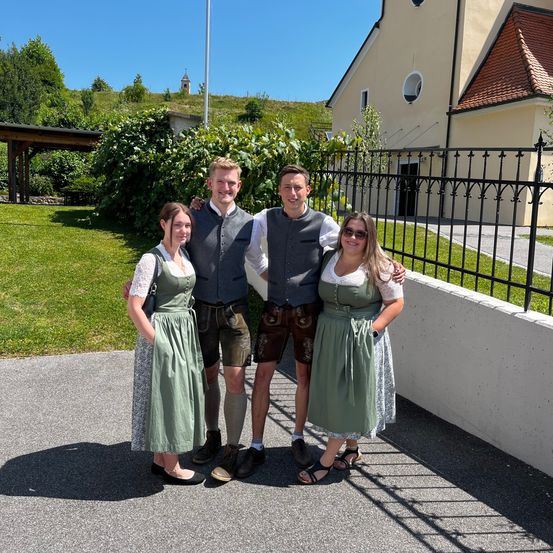 Vier Personen in traditioneller deutscher Kleidung stehen gemeinsam vor einem Gebäude mit eingezäuntem Hof und einem Pavillon an einem sonnigen Tag.