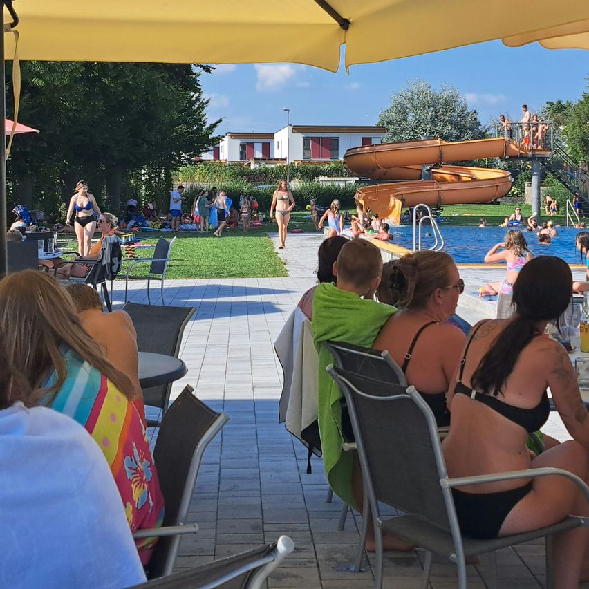 Eine Gruppe von Menschen versammelt sich in einem Wasserpark. Einige Leute schwimmen im Pool, während andere auf Stühlen unter Sonnenschirmen sitzen. Eine Frau geht auf dem Gras.