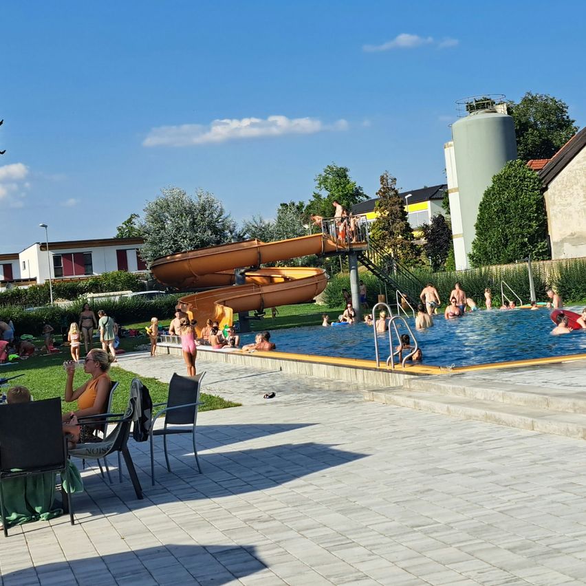 Ein sonniger Tag in einem Freibad. Viele Menschen schwimmen in einem großen Pool, während andere die Rutsche genießen. In der Nähe trinkt eine Frau aus einem Glas, während sie auf einem Stuhl sitzt.