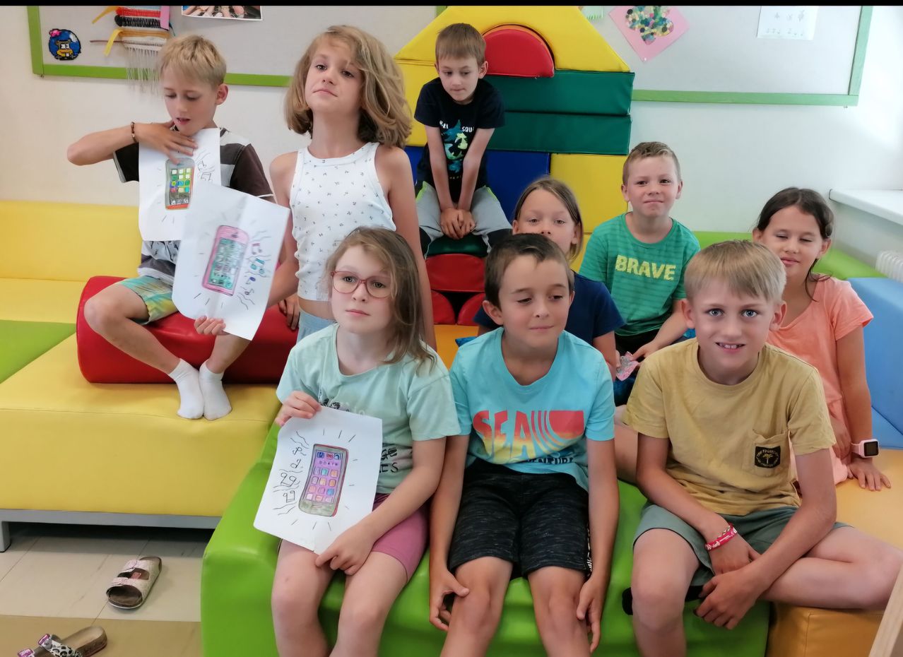 Eine Gruppe von Kindern sitzt zusammen in einem Spielzimmer, einige halten Zeichnungen von Telefonen. Ein Junge auf der linken Seite hält eine Zeichnung mit einem Telefon darauf. Ein anderer Junge hinter ihm steht.