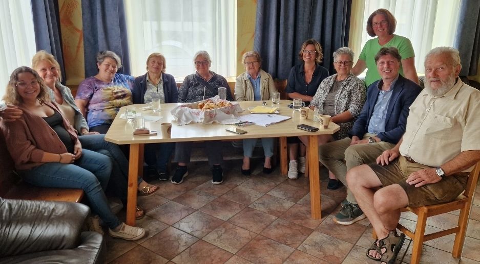 Gruppe von Menschen, die um einen Tisch sitzen, lächeln, mit Essen und Getränken auf dem Tisch und Fenstern mit Vorhängen im Hintergrund.