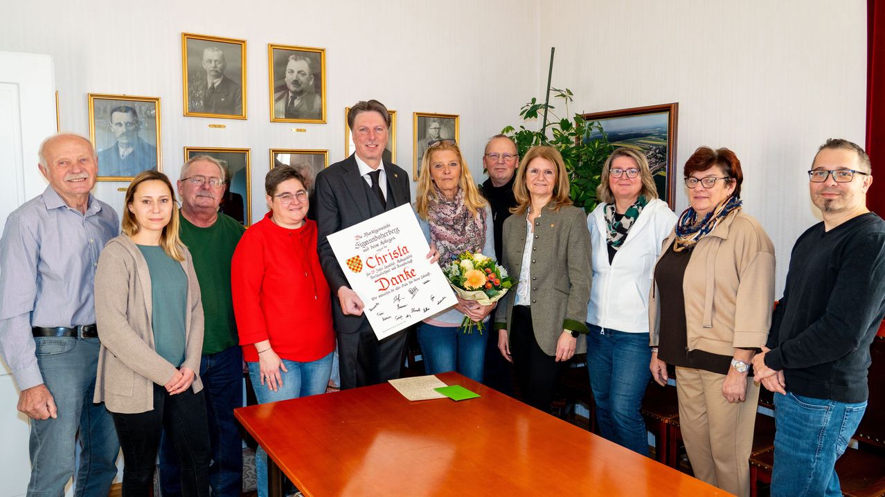 Eine Gruppe von Menschen steht um einen Tisch mit einem großen Zertifikat, eine Person hält es mit einem Blumenstrauß. Sie sind in formeller Kleidung gekleidet. Hinter ihnen schmücken gerahmte Bilder die Wand.