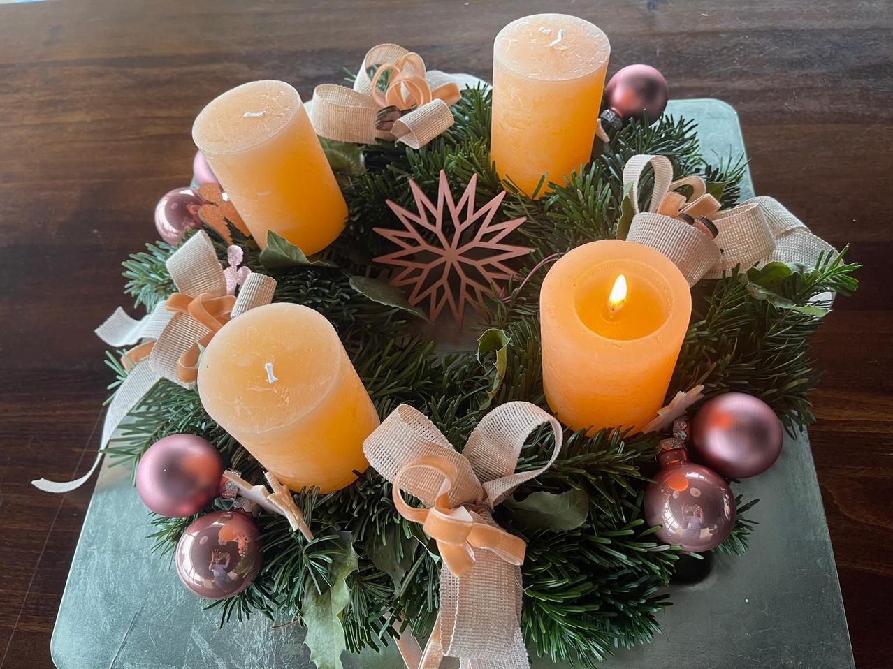 Ein Kranz mit vier gelben Kerzen und roten Kugeln wird auf einer Holzoberfläche präsentiert. Der Kranz besteht aus grünen Tannennadeln und ist mit Bändern und einem Schneeflockenmuster verziert.