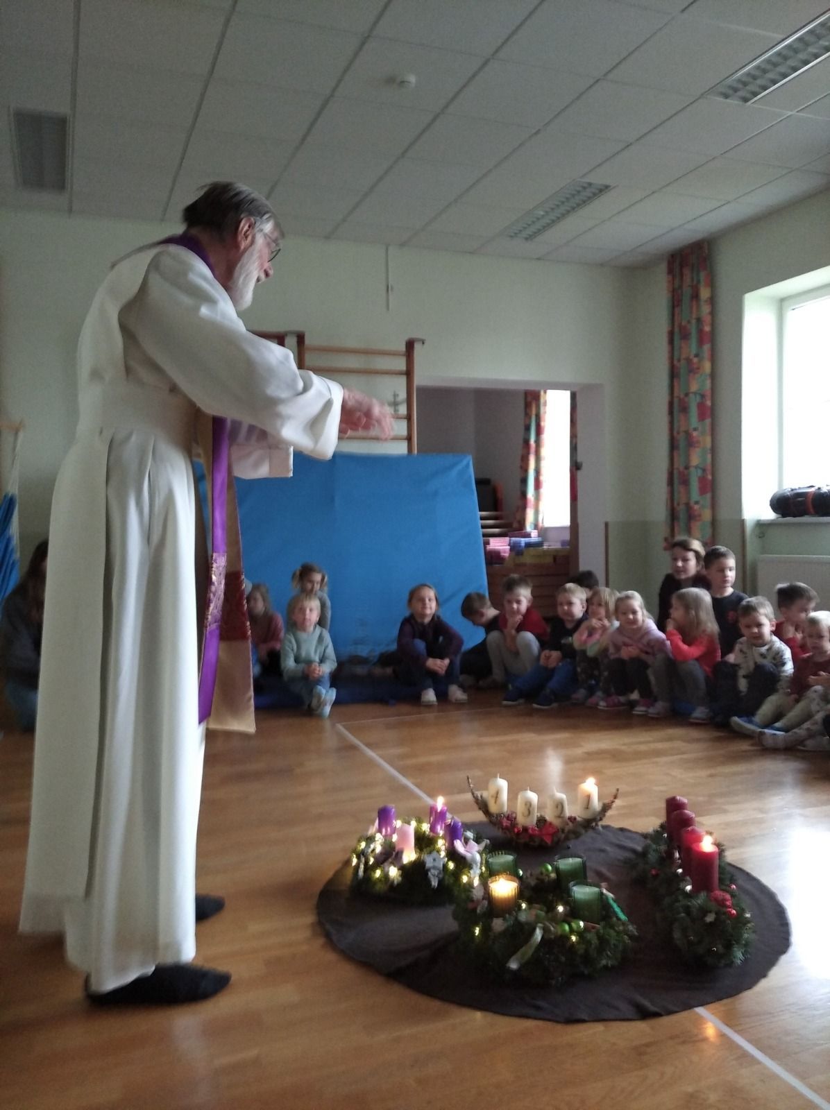 Ein älterer Mann in einer weißen Robe steht vor einer Gruppe von Kindern und hält einen Stab. Sie sitzen um einen Kreis von brennenden Kerzen herum auf dem Boden.