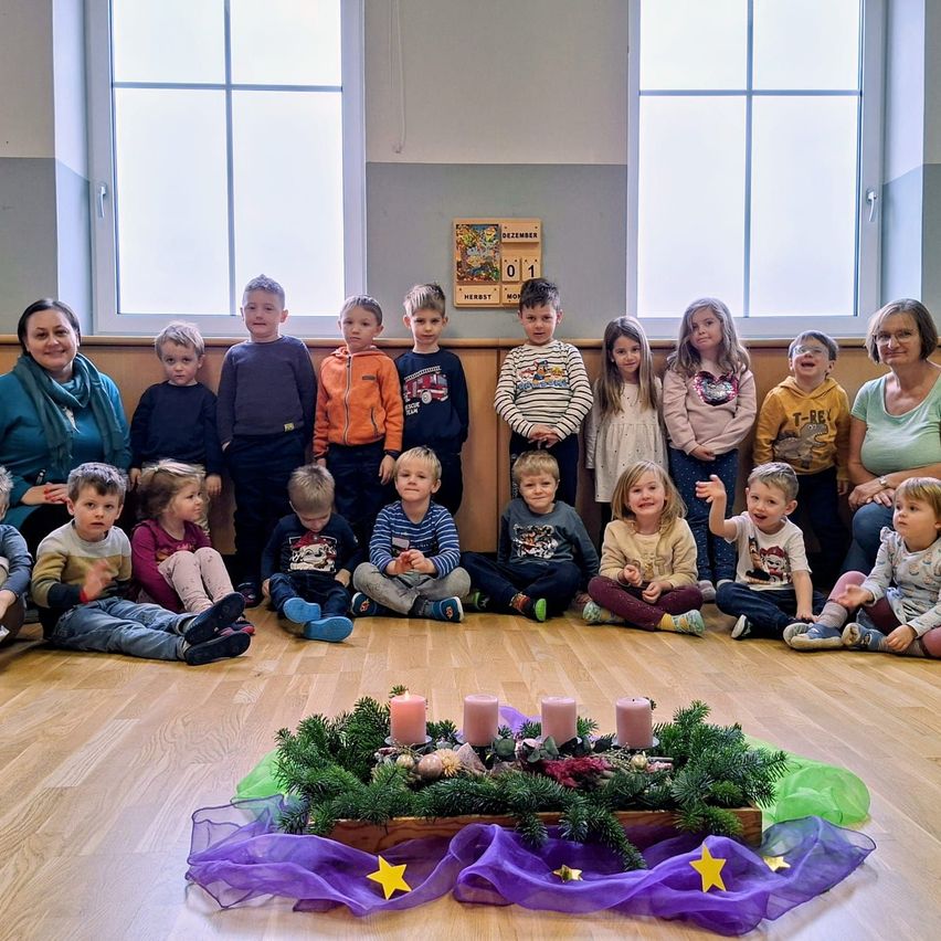 Gruppenfoto von Kindern und zwei Frauen, die auf dem Boden sitzen, umgeben von einem Kranz mit Kerzen.