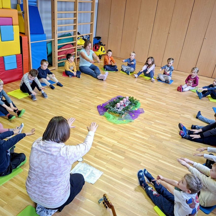 Eine Frau leitet einen Kreis junger Kinder in einem Raum, wahrscheinlich einem Kindergarten. Die Kinder sitzen auf bunten Matten, und eine zentrale Dekoration ist sichtbar.