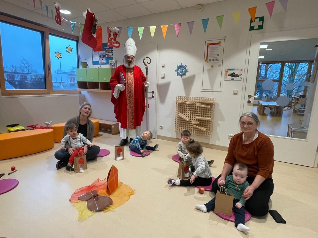 Ein Priester in roter Kleidung hält einen Stab und spricht zu einer Gruppe sitzender Kinder und Erwachsener in einem Raum mit bunten Dekorationen und einer Spielstruktur.