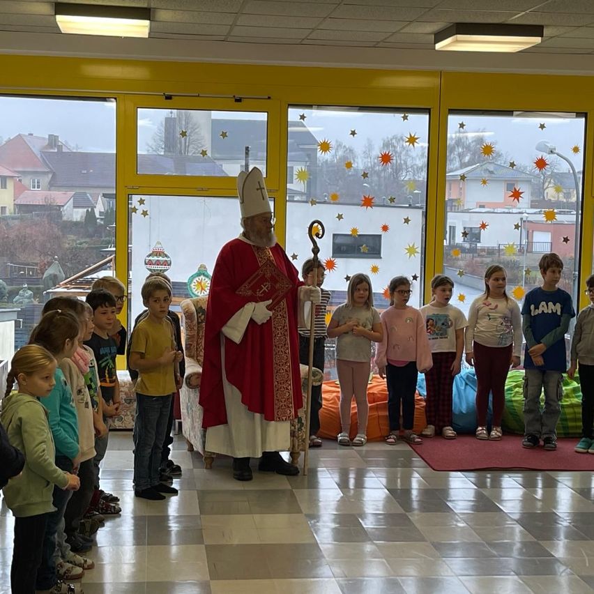 Ein Priester in einer roten Robe steht in einem Klassenzimmer mit Kindern. Das Klassenzimmer hat gelbe Wände und Glastüren mit Sternen. Die Kinder stehen vor dem Priester.