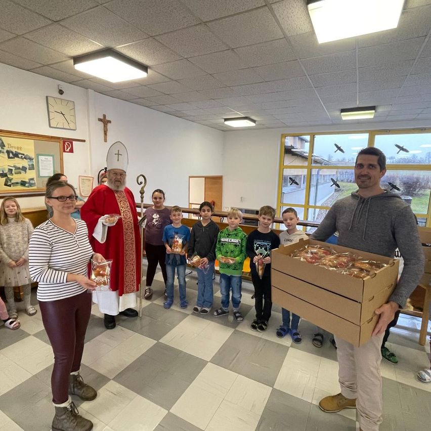 Ein Priester in roten Roben hält eine Box, während ein Mann in einer grauen Kapuzenjacke eine Box mit Essen hält. Sie sind von Kindern umgeben, die ihre eigenen Essensboxen halten.
