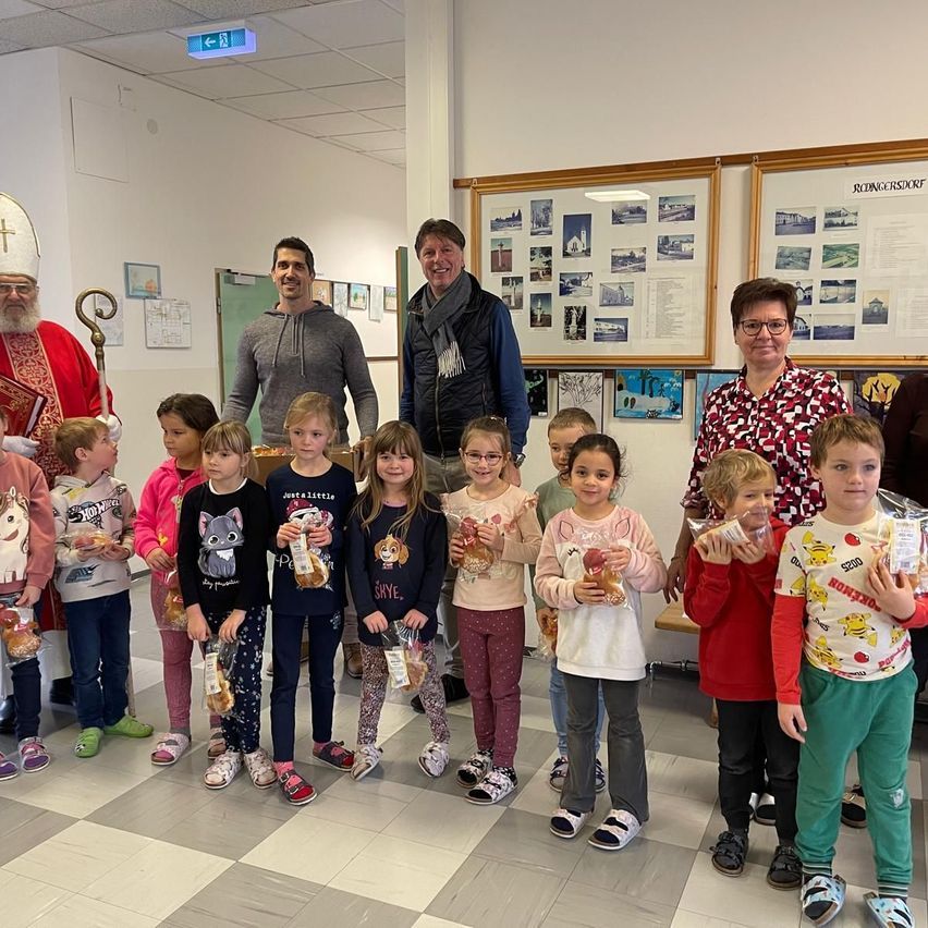 Eine Gruppe von Kindern und Erwachsenen, einschließlich eines Priesters, steht in einem Raum mit einem gemusterten Boden. Die Kinder halten Snacks in Plastiktüten, und die Erwachsenen lächeln. Es gibt Bilderrahmen an der Wand und eine weiße Decke mit Lichtern.