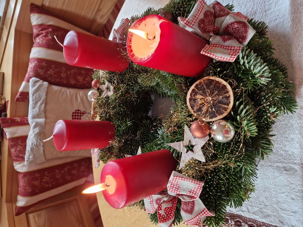 Ein Kranz mit vier roten Kerzen, geschmückt mit roten Bändern und einer Orangenscheibe, steht auf einem Holztisch inmitten einer festlichen Atmosphäre.