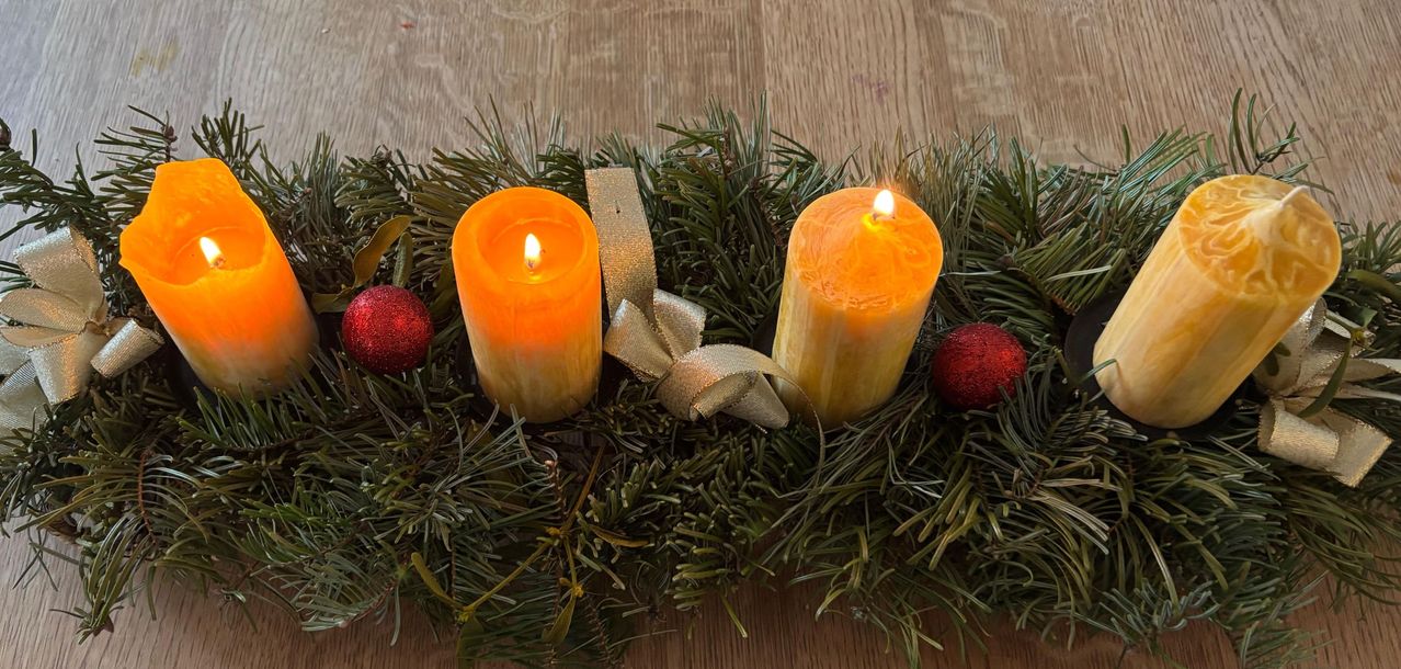 Zwei gelbe Kerzen mit Schleifen und roten Kugeln auf einem Kranz aus grünen Kiefernnadeln auf einer hölzernen Oberfläche.