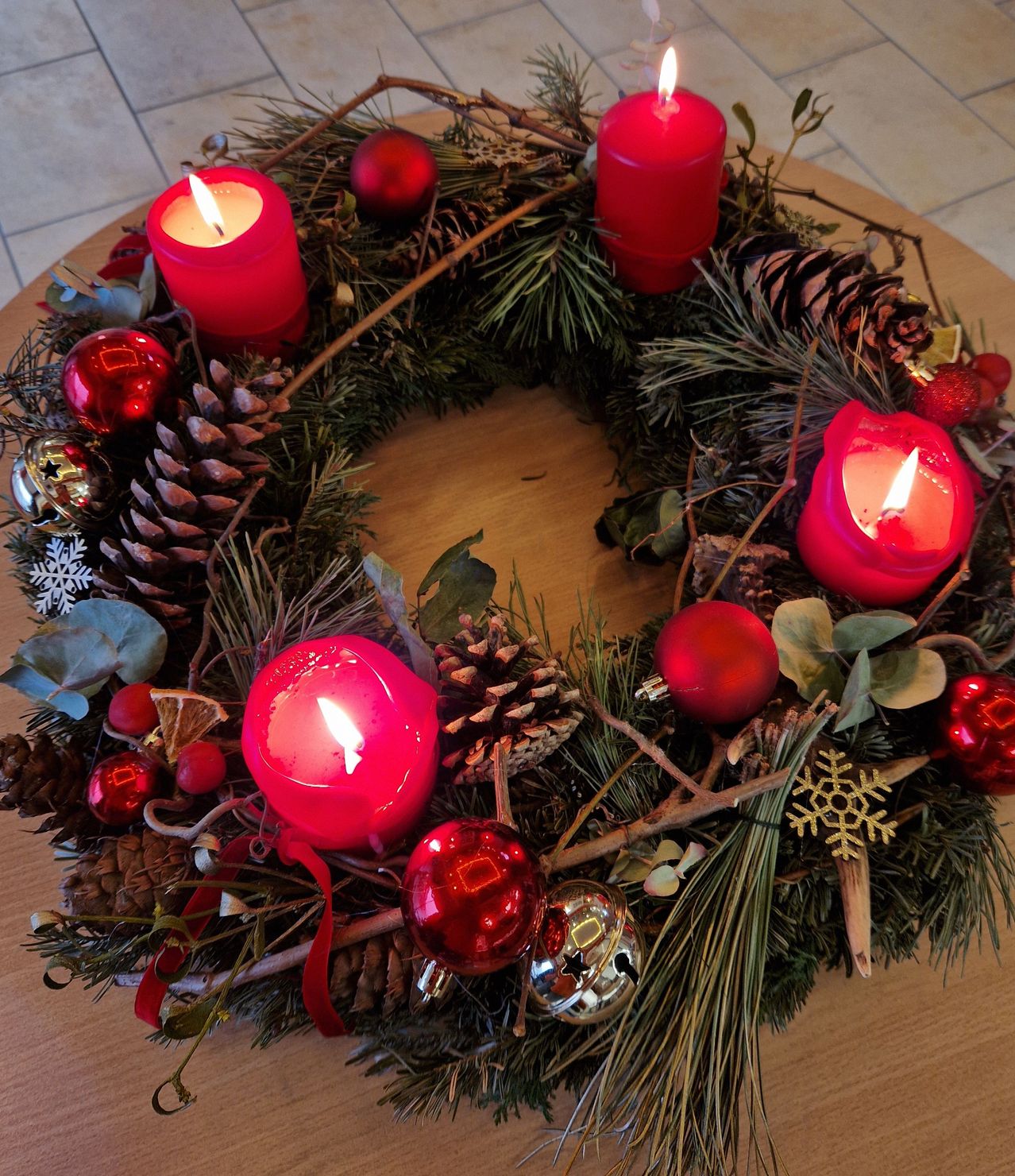 Ein Kranz auf einem Holztisch ist mit roten Ornamenten, Tannenzapfen und vier brennenden roten Kerzen geschmückt. Es gibt auch rote Kugeln und silberne Glocken, wobei Grünpflanzen und Stechpalmenblätter einen festlichen Akzent setzen.