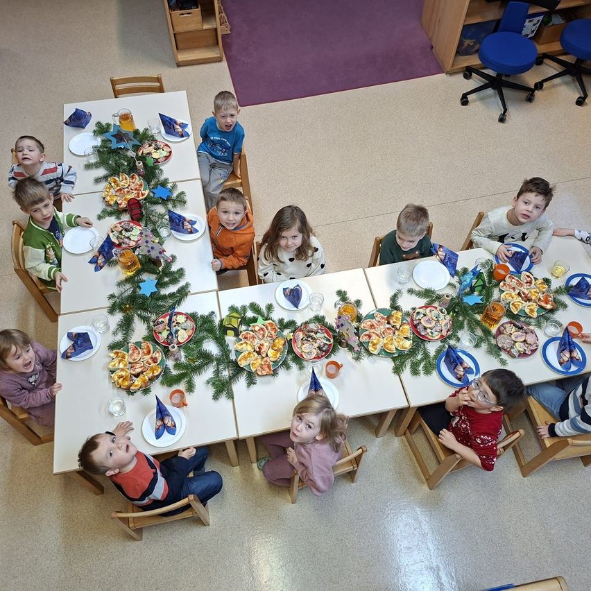 Eine Gruppe junger Kinder sitzt um einen langen Tisch, der mit Essen gefüllt ist. Der Tisch ist mit Girlanden und Papiersternen geschmückt. Die Kinder schauen in die Kamera.