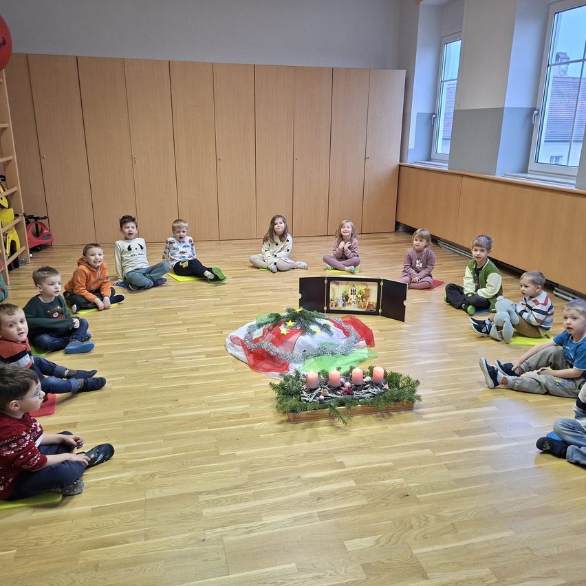 Eine Gruppe von Kindern sitzt im Kreis auf dem Boden. Vor ihnen steht eine Weihnachtsdekoration mit Kerzen. Der Raum hat Holzböden und -wände.