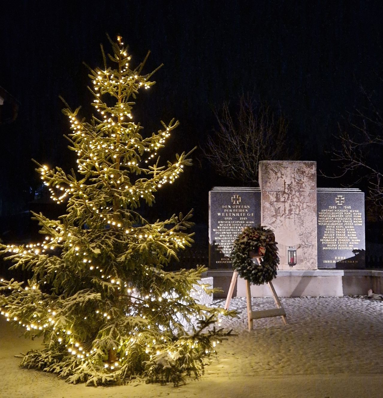 Ein beleuchteter Weihnachtsbaum steht in der Nähe eines Denkmals mit einem Kranz davor. Das Denkmal hat eingravierte Texte und ein Kreuzsymbol darauf. Der Boden ist schneebedeckt.