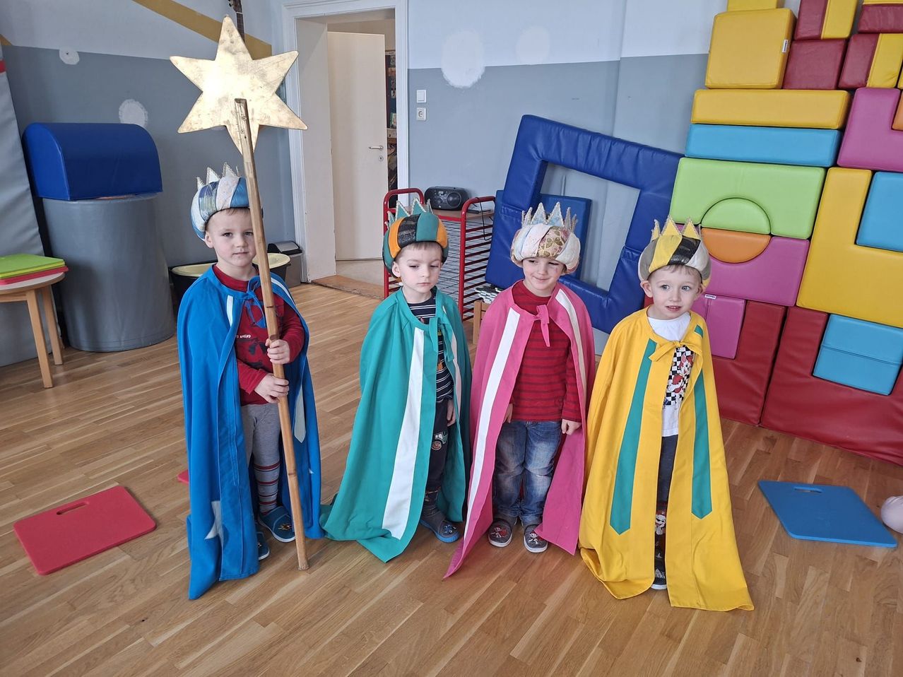 Vier junge Jungen, die als Könige gekleidet sind, stehen in einem Raum mit Holzböden und tragen bunte Umhänge und Kronen. Einer hält einen Sternenstab. Dahinter befinden sich bunte Kissen und ein blaues Objekt.