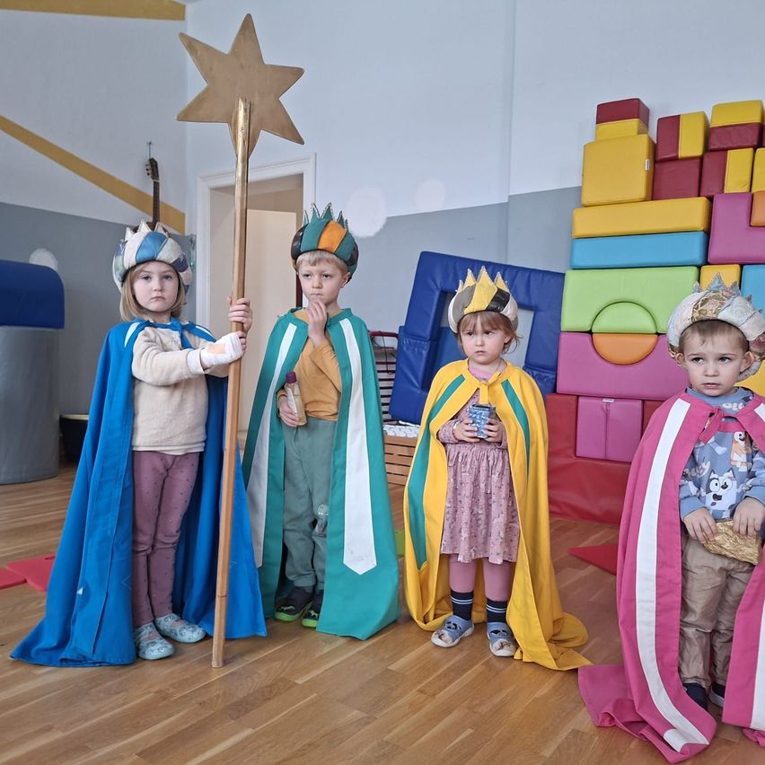 Fünf kleine Kinder in bunten Kostümen stehen in einem Raum. Eines hält einen sternenbesetzten Stab, während die anderen Kronen tragen. Dahinter sind bunte Bausteine gestapelt.