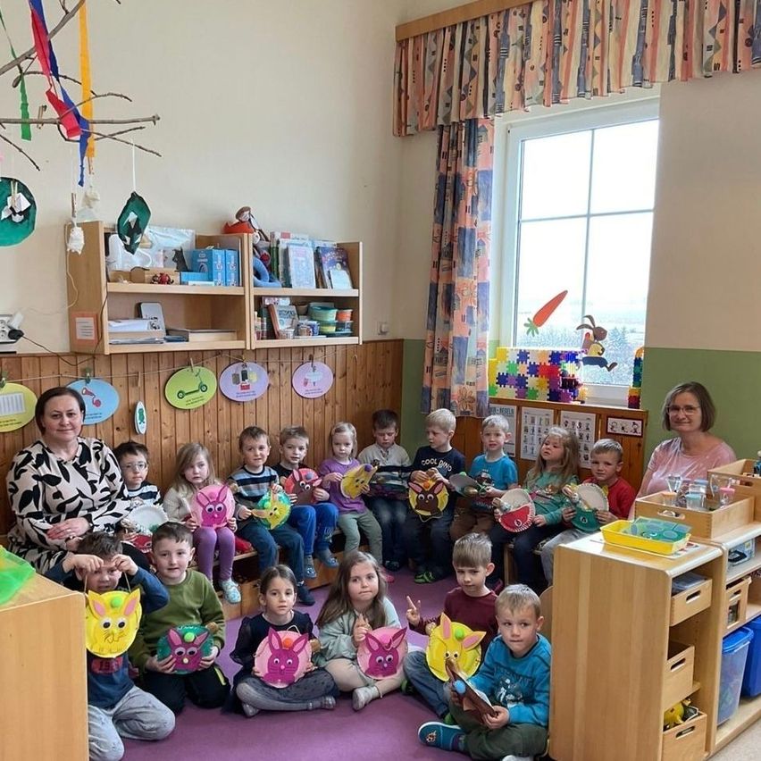 A group of children and two teachers in a classroom, sitting on the floor and holding paper crafts. The classroom has wooden shelves, curtains, and a window with a rabbit figure.