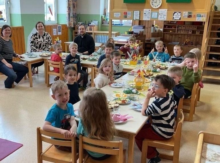 A classroom with children seated around tables, eating. A woman in a black and white dress sits in the back. Adults stand behind the tables.