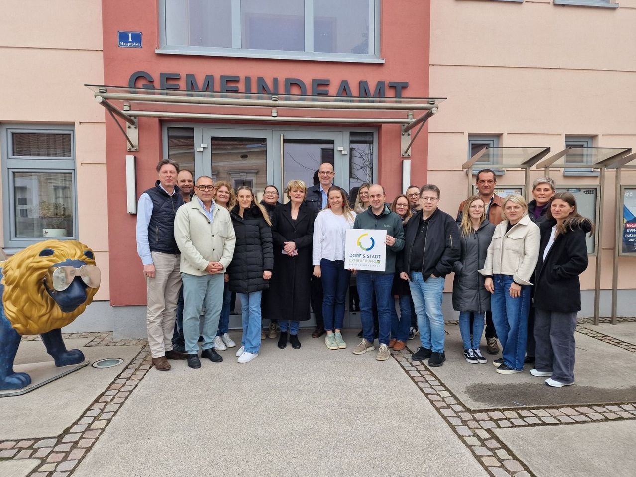 A group of people stands in front of a building with a sign that says Gemeindeamt. They are all dressed in winter clothes and are posing for a photo.