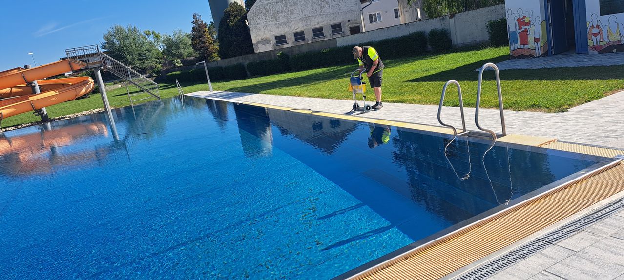 Ein Mann in einem reflektierenden Vest reinigt einen großen Pool an einem sonnigen Tag mit einem Gebäude im Hintergrund.