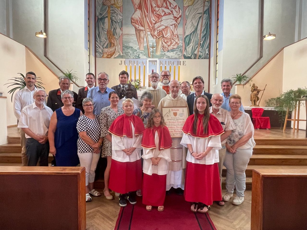 Eine Gruppe von Menschen in traditioneller Kleidung steht in einer Kirche, wobei ein Priester ein gerahmtes Zertifikat in der Hand hält. Dahinter ziert eine große Wandmalerei die Wand.
