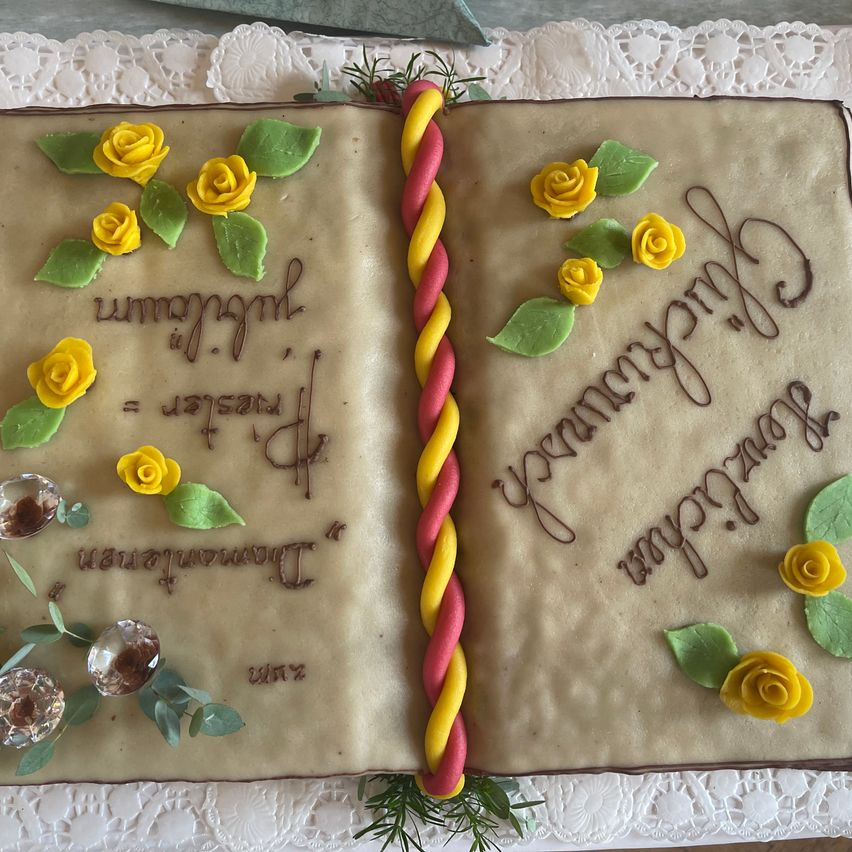 Ein Kuchen in Form eines offenen Buches mit gelben Rosen und grünen Blättern auf den Seiten, und der Text 'Happy Birthday' in brauner Glasur geschrieben.