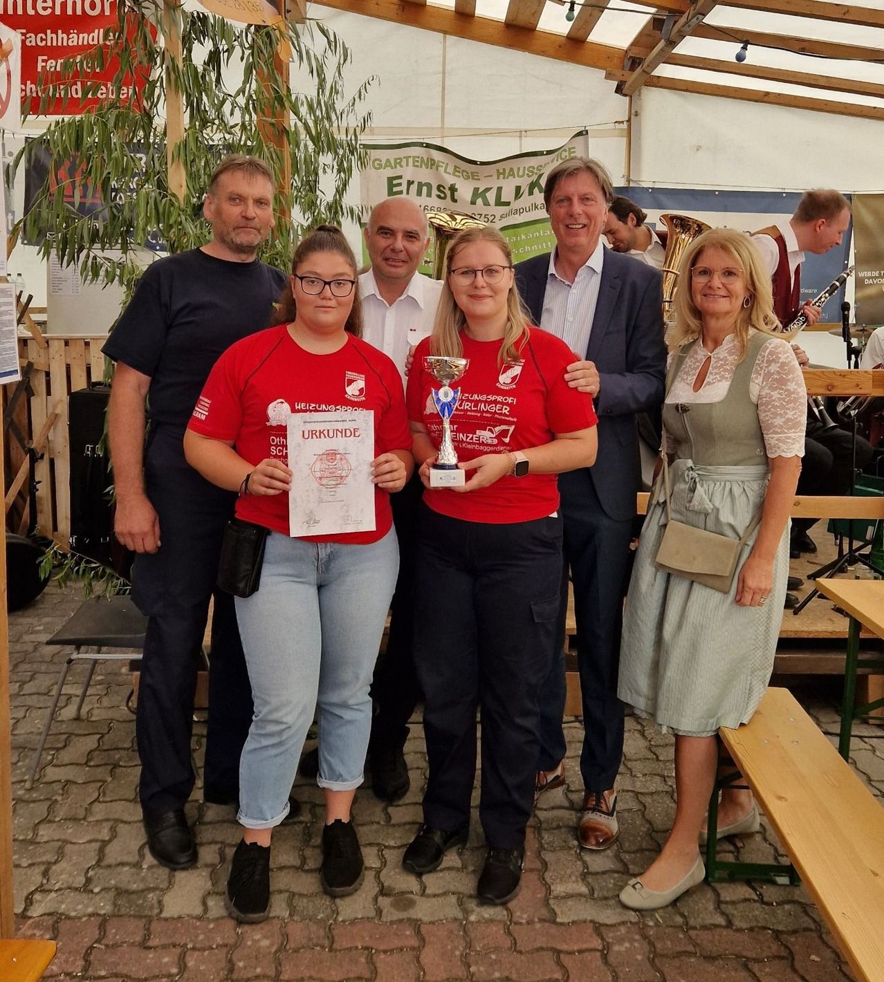 Five people stand together, two women holding trophies and certificates, with a man in a suit and two men in casual attire. They are in an outdoor setting with a tent and wooden benches.